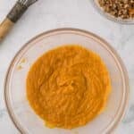 A glass bowl containing orange pumpkin puree, with a wire whisk and a separate bowl of pecan crumble nearby.