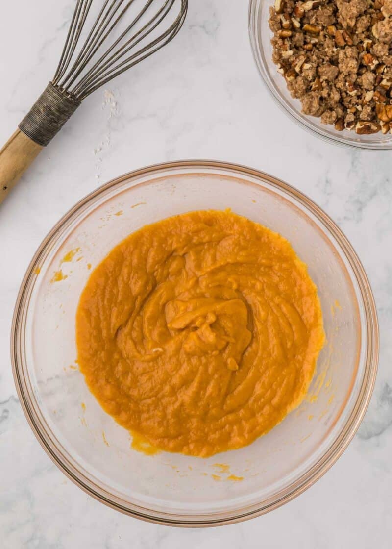 A glass bowl containing orange pumpkin puree, with a wire whisk and a separate bowl of pecan crumble nearby.