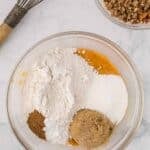 A glass bowl containing flour, brown sugar, white sugar, and spices atop a pumpkin mixture, with a whisk and a bowl of pecan topping nearby.