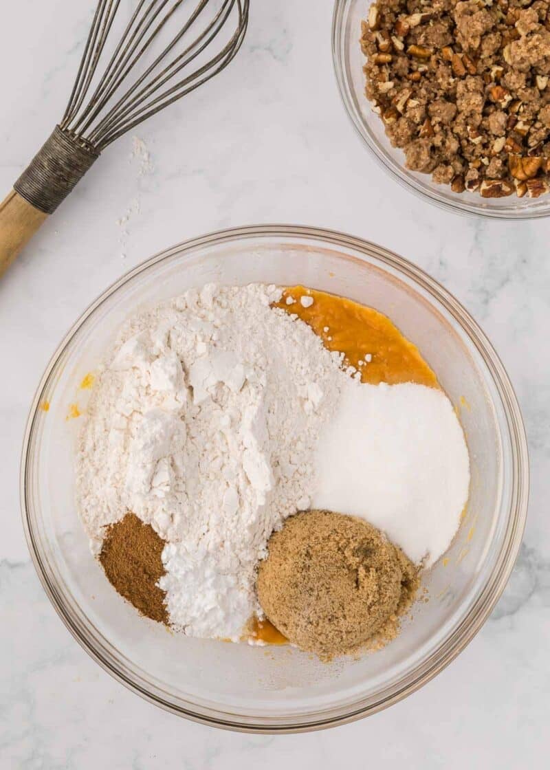 A glass bowl containing flour, brown sugar, white sugar, and spices atop a pumpkin mixture, with a whisk and a bowl of pecan topping nearby.