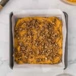 Pumpkin batter topped with a pecan crumble in a square baking pan lined with parchment paper.