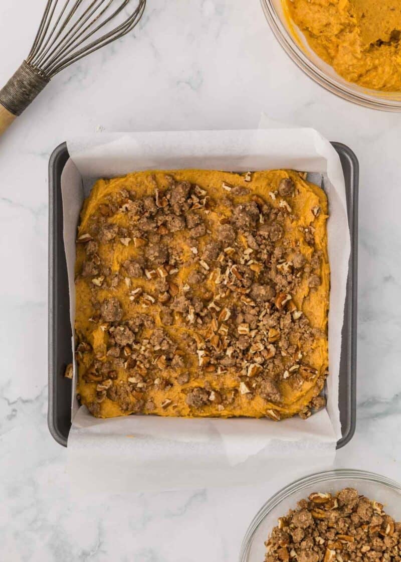 Pumpkin batter topped with a pecan crumble in a square baking pan lined with parchment paper.