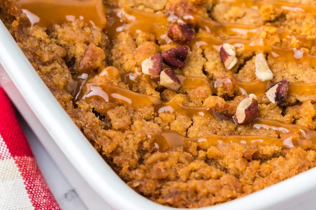 A square baking dish containing a pumpkin dump cake drizzled with caramel sauce and sprinkled with pecan pieces.