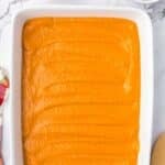 A white baking dish filled with smooth orange pumpkin filling, with small bowls of melted butter and crumbled crust nearby.