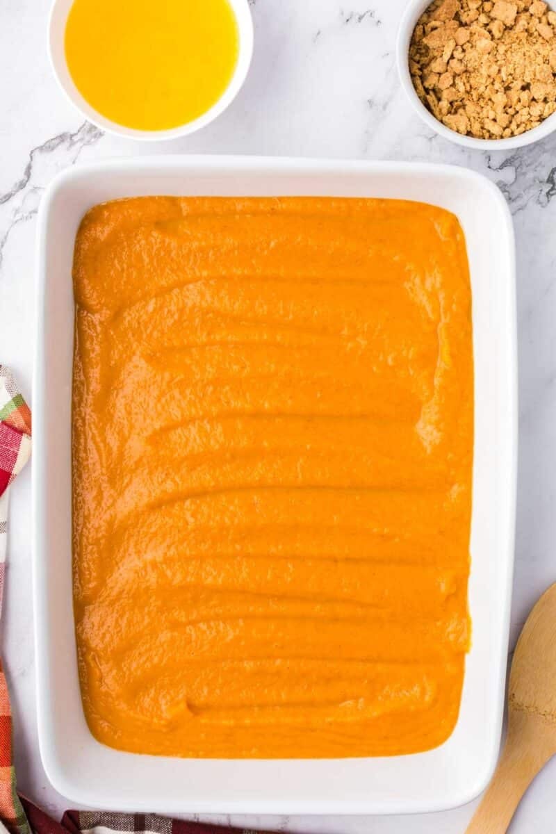 A white baking dish filled with smooth orange pumpkin filling, with small bowls of melted butter and crumbled crust nearby.