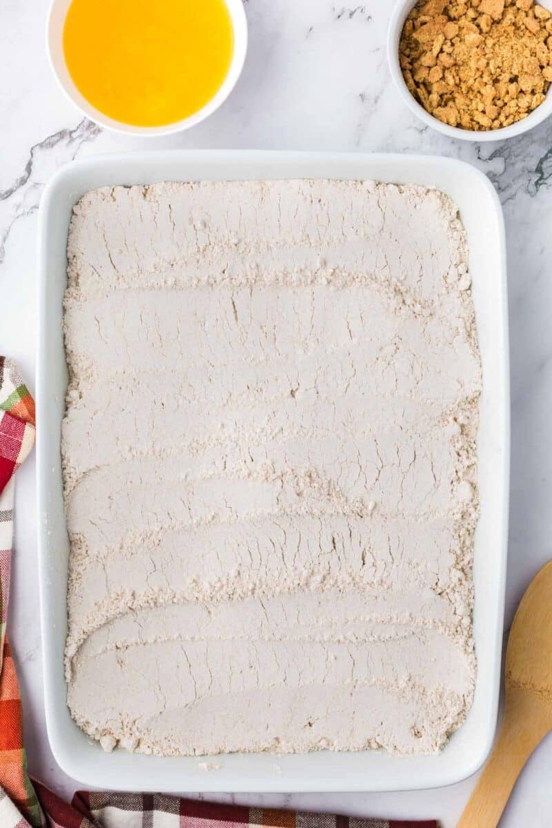 A rectangular baking dish filled with a layer of dry flour or cake mix, accompanied by small bowls of melted butter and crumbled topping.