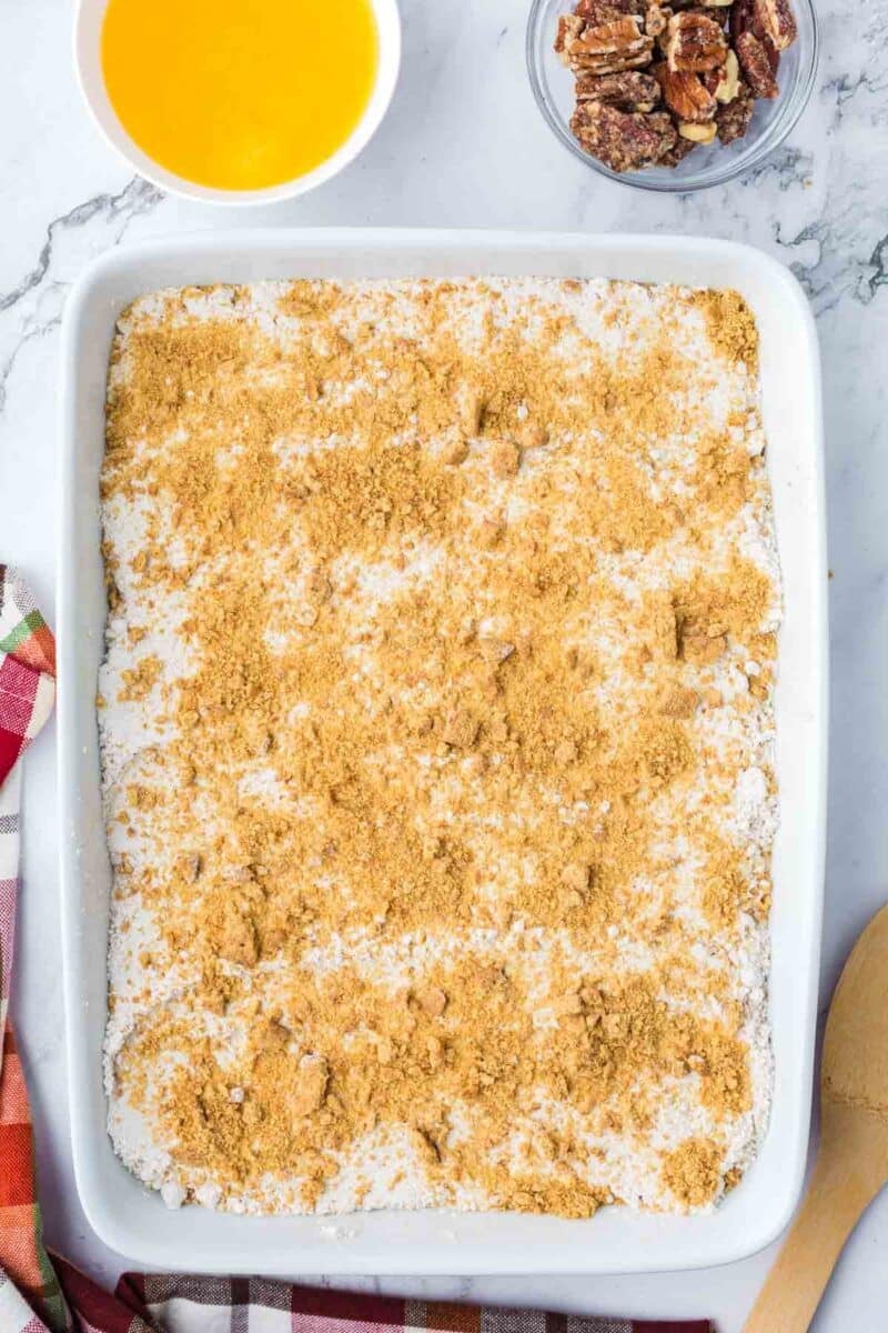 A rectangular baking dish filled with a pumpkin dump cake, alongside small bowls of melted butter and pecans.