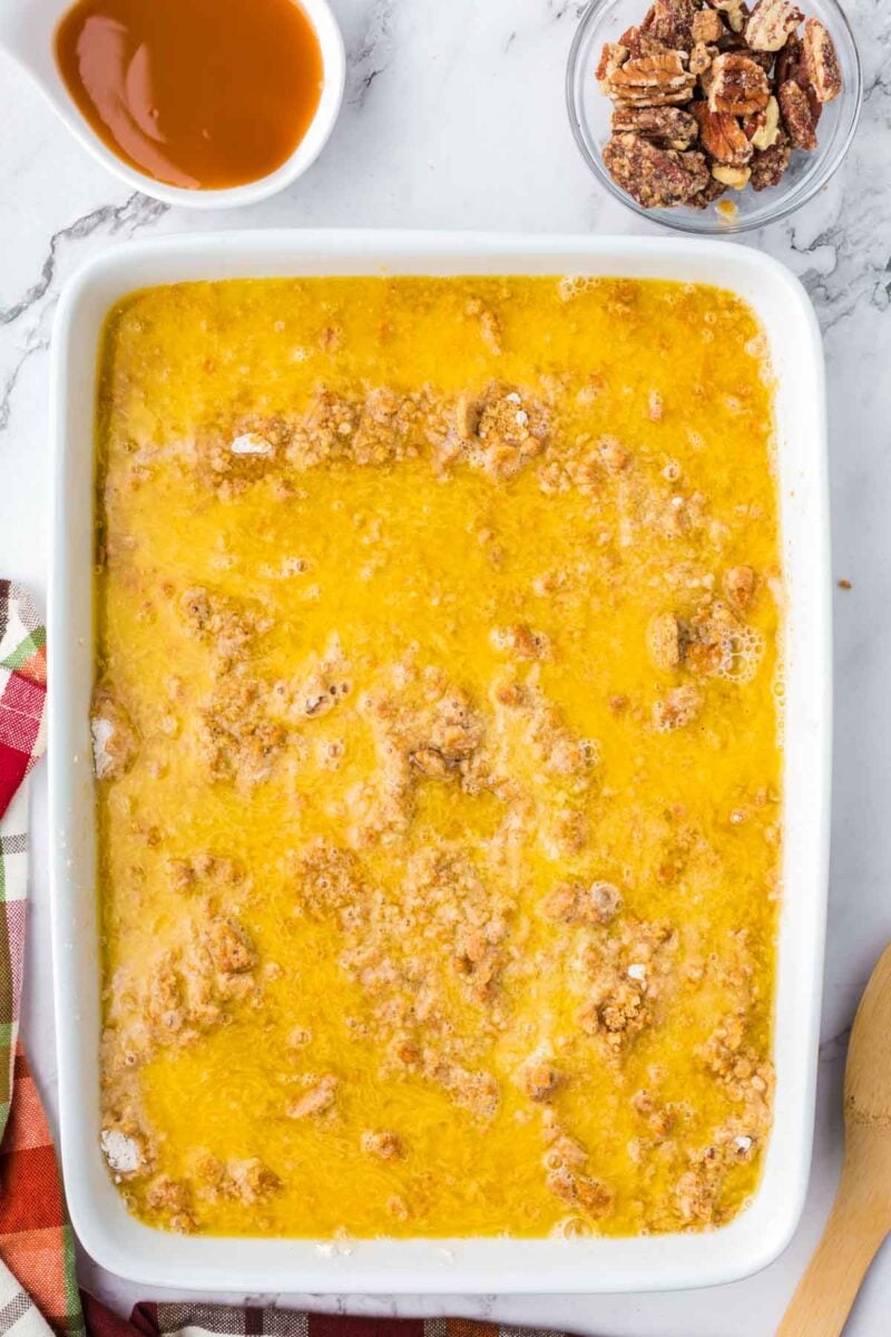A rectangular baking dish filled with a golden butter and crumb mixture, accompanied by small bowls of caramel sauce and pecans.