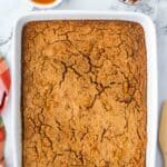 A rectangular baked pumpkin dump cake in a white dish, with small bowls of caramel sauce and chopped pecans nearby.