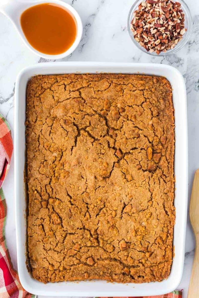 A rectangular baked pumpkin dump cake in a white dish, with small bowls of caramel sauce and chopped pecans nearby.