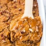A wooden spatula lifts a square of pecan-topped pumpkin dump cake drizzled with caramel sauce from a baking dish.