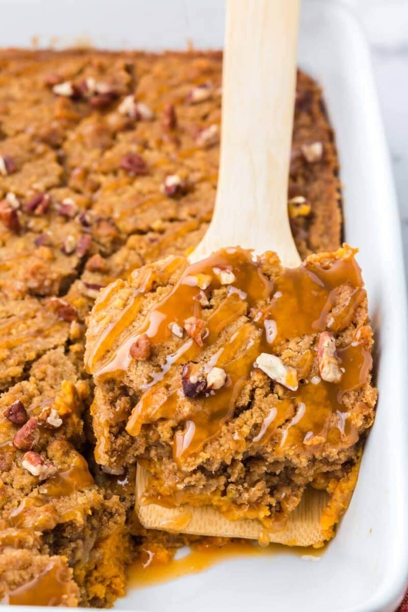 A wooden spatula lifts a square of pecan-topped pumpkin dump cake drizzled with caramel sauce from a baking dish.
