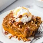 A slice of pumpkin dump cake topped with whipped cream, caramel sauce, and pecans on a white plate with a fork.