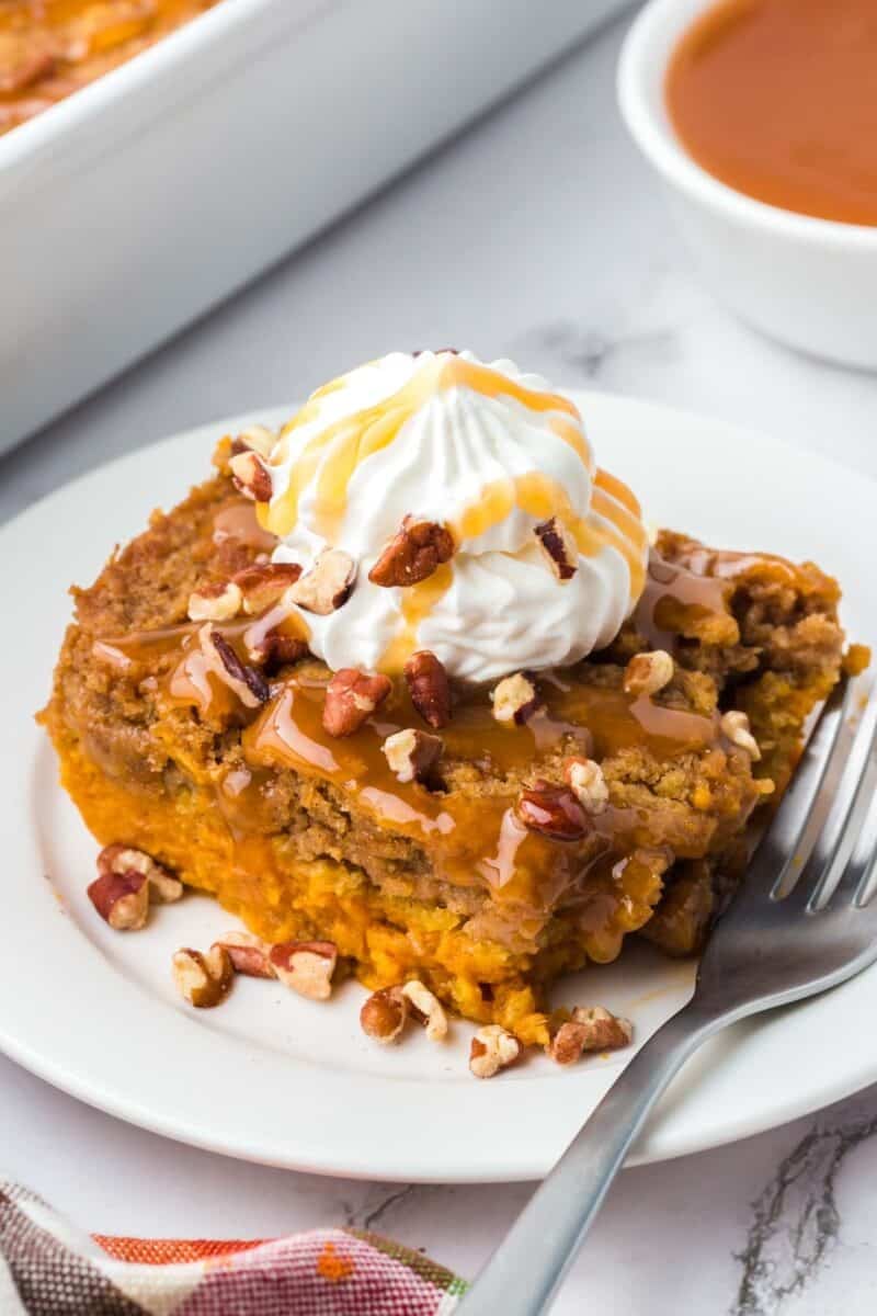 A slice of pumpkin dump cake topped with whipped cream, caramel sauce, and pecans on a white plate with a fork.