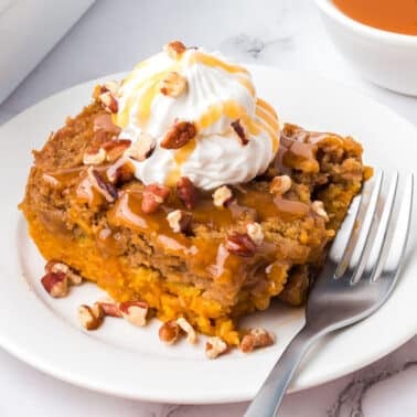 A square piece of pumpkin dump cake&nbsp;topped with whipped cream, caramel sauce, and pecans on a white plate with a fork.
