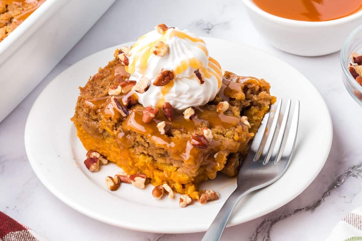 A square piece of pumpkin dump cake topped with whipped cream, caramel sauce, and pecans on a white plate with a fork.