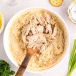 A wooden spoon mixing shredded chicken into a creamy dip in a white bowl, surrounded by crackers, herbs, and spices.