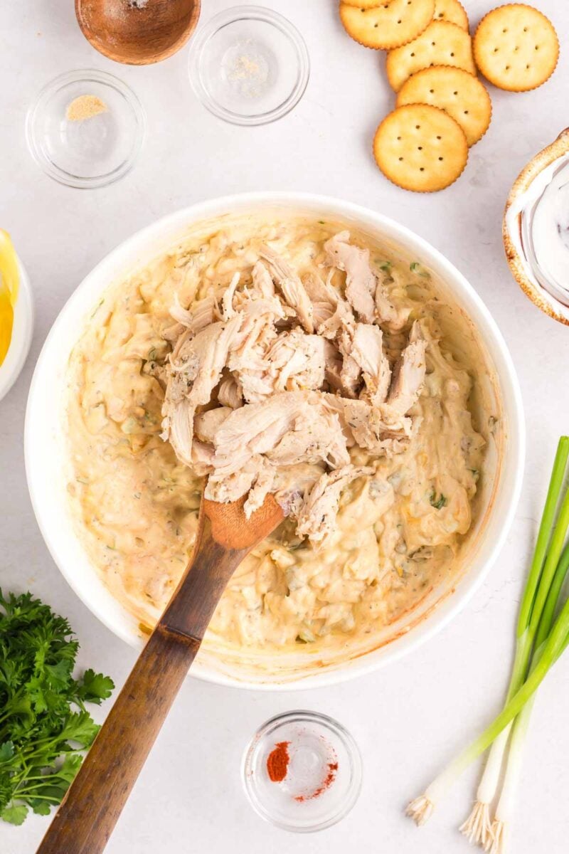 A wooden spoon mixing shredded chicken into a creamy dip in a white bowl, surrounded by crackers, herbs, and spices.