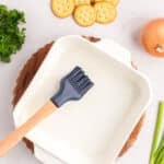 A pastry brush sits in a white square baking dish, surrounded by crackers, parsley, an onion, and green onions.