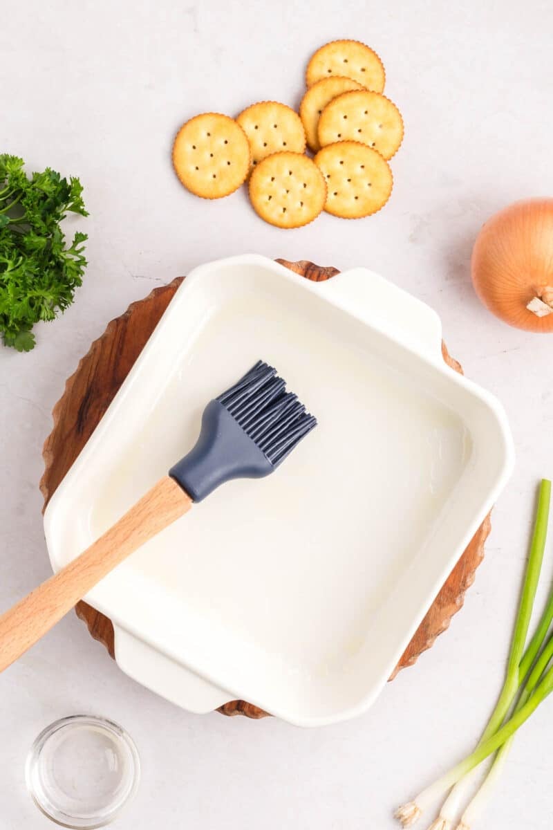 A pastry brush sits in a white square baking dish, surrounded by crackers, parsley, an onion, and green onions.