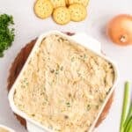A square white baking dish filled with ritz cracker chicken casserole, served with round crackers, a whole onion, green onions, and parsley.
