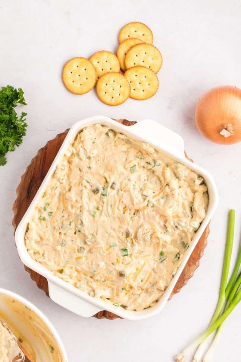 A square white baking dish filled with ritz cracker chicken casserole, served with round crackers, a whole onion, green onions, and parsley.