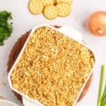 A square casserole dish topped with a golden-brown cracker crumb crust, surrounded by whole crackers, an onion, green onions, and parsley.