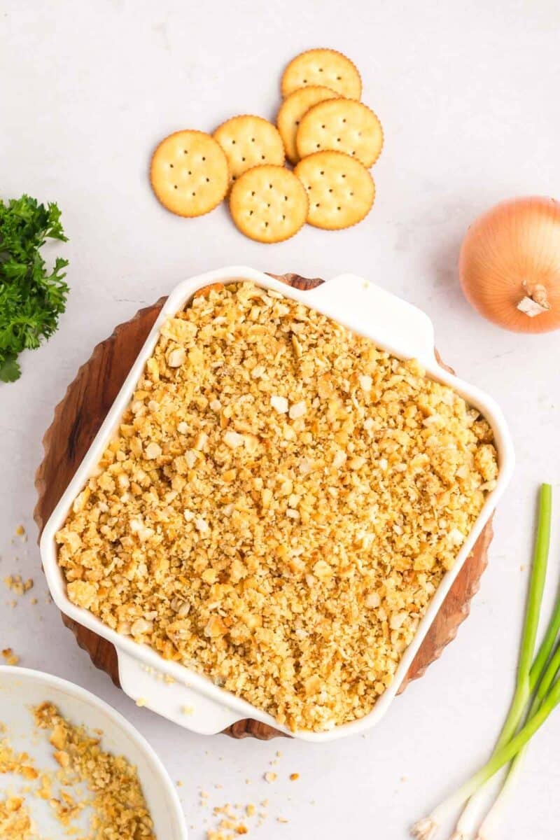 A square casserole dish topped with a golden-brown cracker crumb crust, surrounded by whole crackers, an onion, green onions, and parsley.