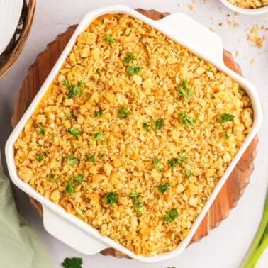 A square white baking dish filled with a ritz cracker chicken casserole topped with golden breadcrumbs and garnished with parsley, served on a wooden board with an onion and green onions.