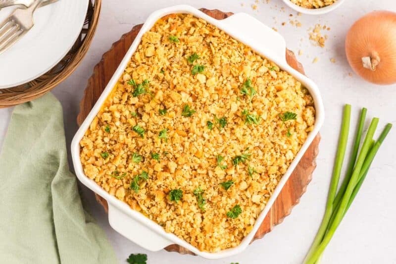 A square white baking dish filled with a ritz cracker chicken casserole topped with golden breadcrumbs and garnished with parsley, served on a wooden board with an onion and green onions.