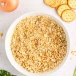 A bowl filled with crushed crackers, surrounded by a whole onion, whole crackers, parsley, and green onions.