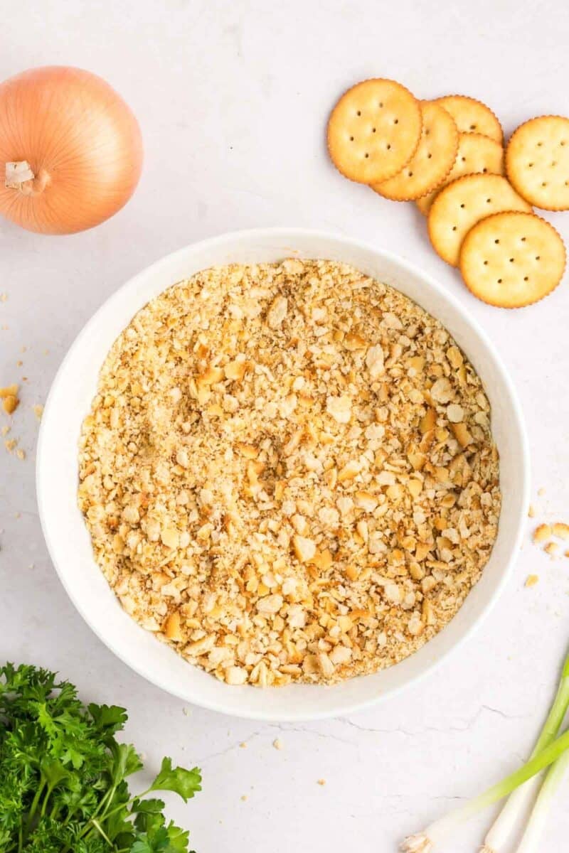 A bowl filled with crushed crackers, surrounded by a whole onion, whole crackers, parsley, and green onions.