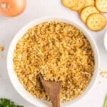 A bowl of crushed crackers with a wooden spoon, surrounded by an onion, whole crackers, and fresh parsley.