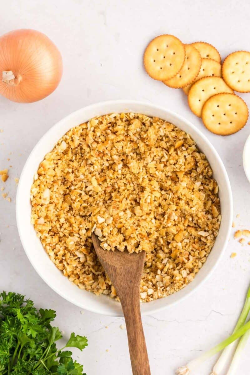 A bowl of crushed crackers with a wooden spoon, surrounded by an onion, whole crackers, and fresh parsley.