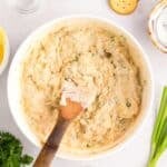 A wooden spoon resting in a bowl of creamy dip, surrounded by crackers, herbs, and small bowls of spices.