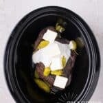 A beef roast in a slow cooker topped with white sauce, butter cubes, and pickled peppers.