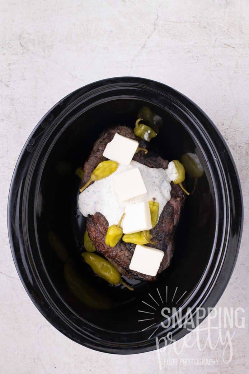 A beef roast in a slow cooker topped with white sauce, butter cubes, and pickled peppers.