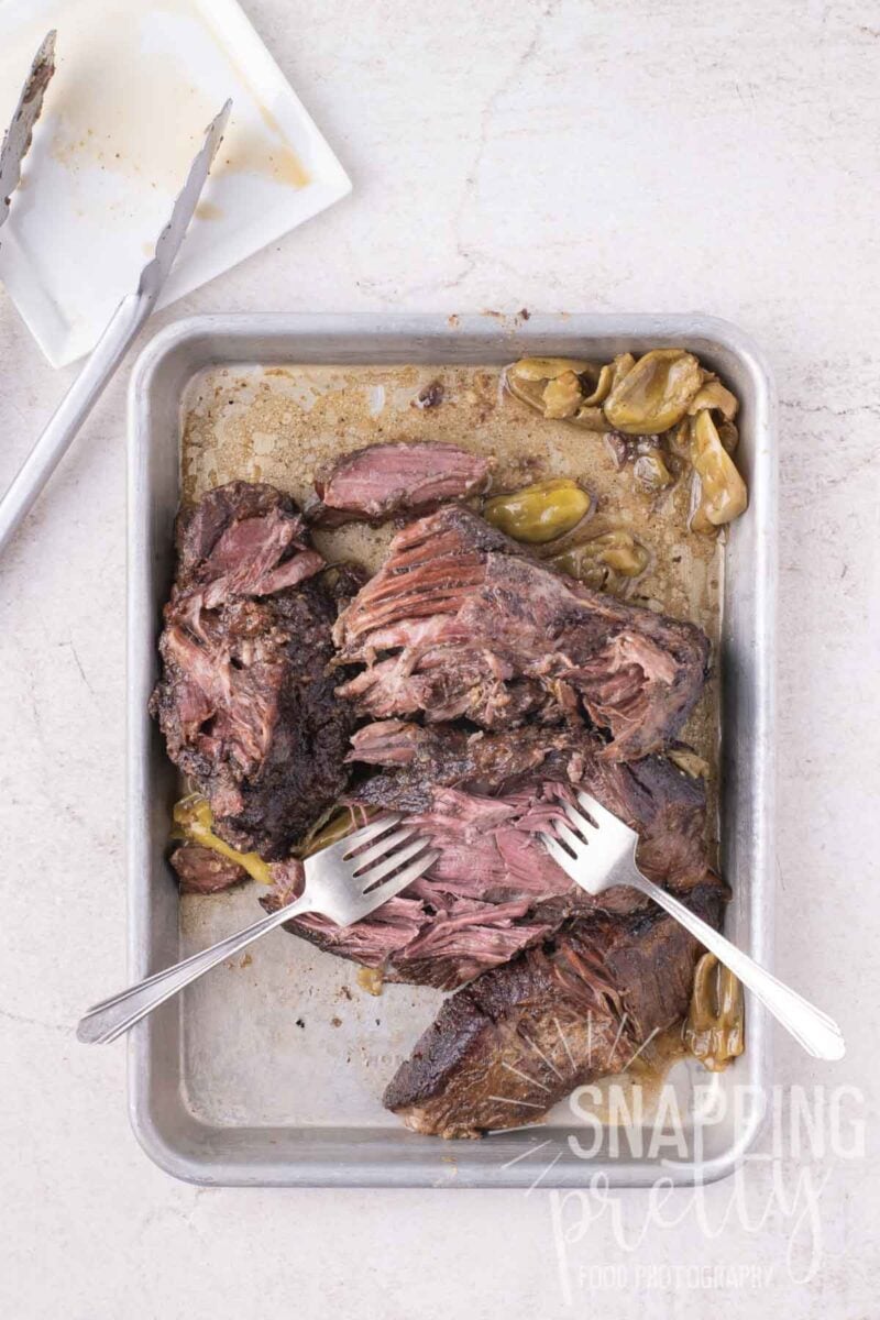 Shredded roast beef with peppers in a metal baking pan being pulled apart with two forks.