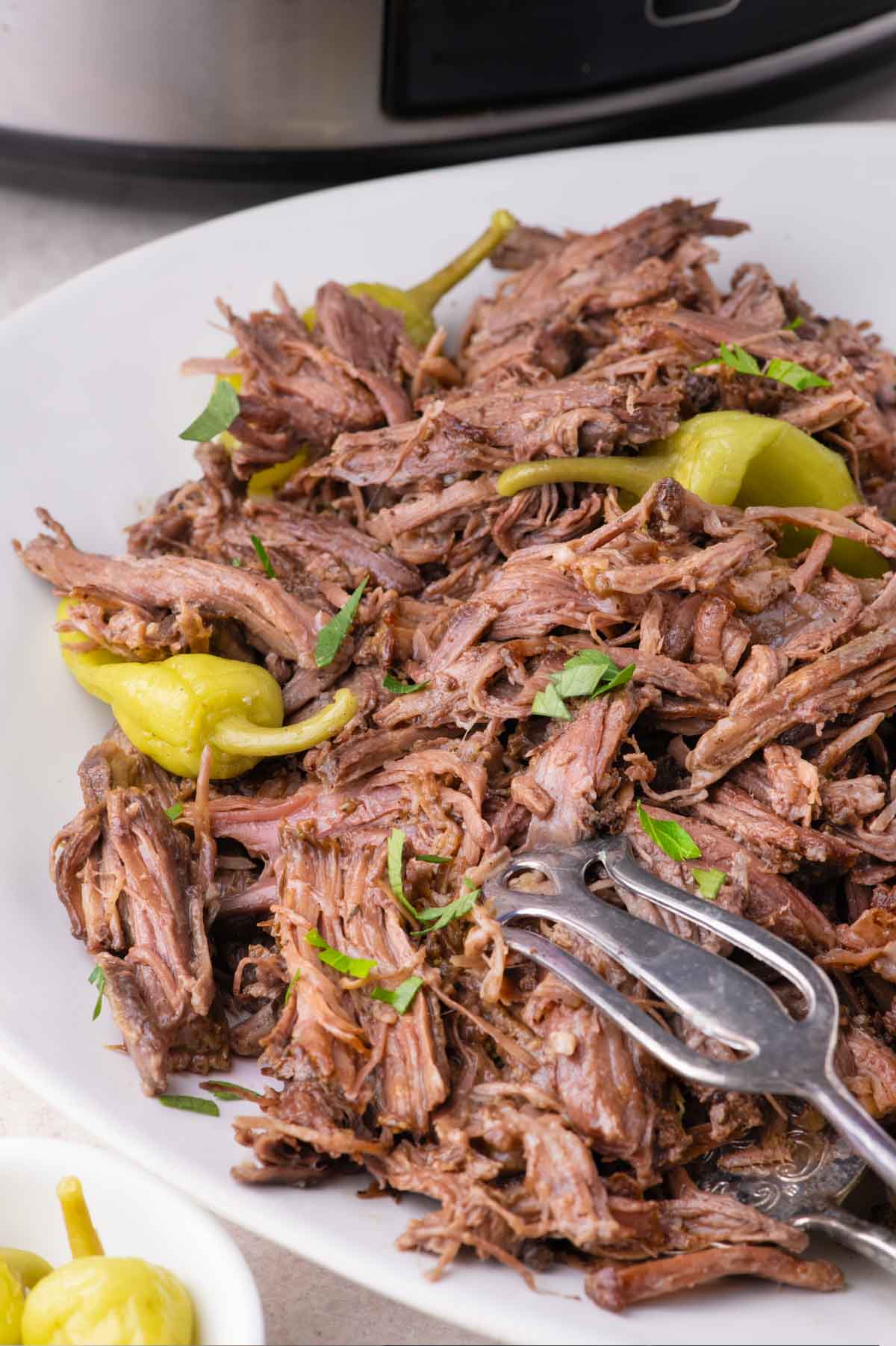 A plate of shredded beef topped with whole pepperoncini peppers and fresh herbs, with a serving fork resting on the side.