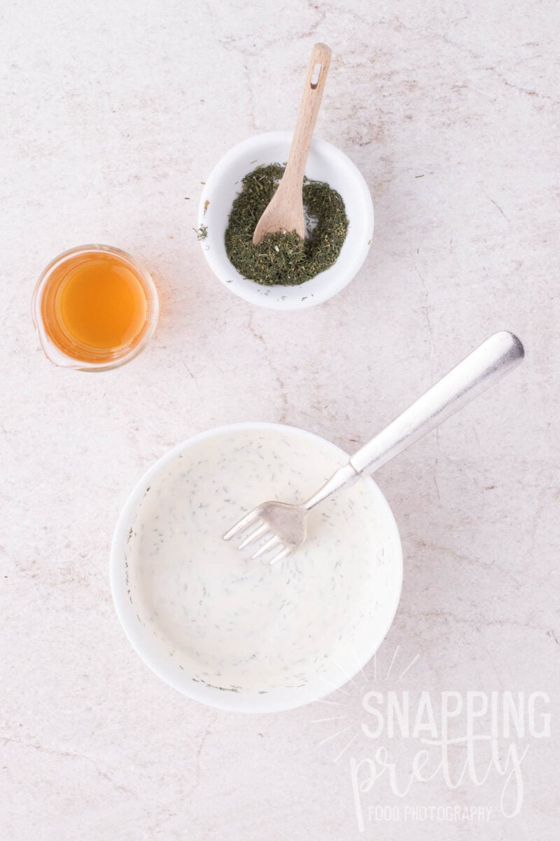 A bowl of creamy herb dressing with a fork, a small bowl of dried herbs with a spoon, and a glass of honey.