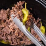 Metal tongs holding a portion of mississippi pot roast and a whole pepper over a slow cooker.