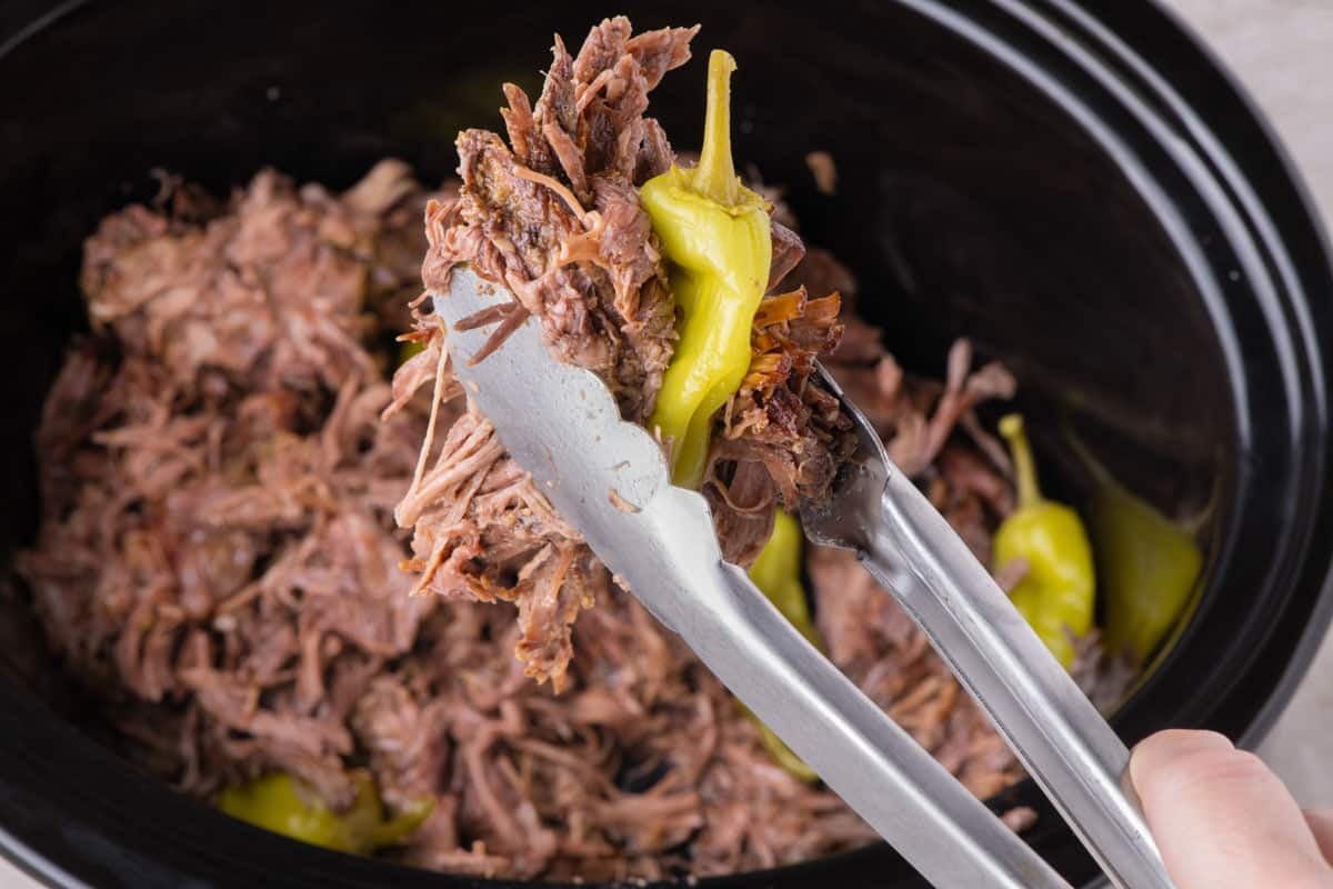 Metal tongs holding a portion of mississippi pot roast and a whole pepper over a slow cooker.