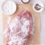 A raw beef roast coated in flour and pepper on parchment paper, with small bowls of flour, salt, and pepper nearby.