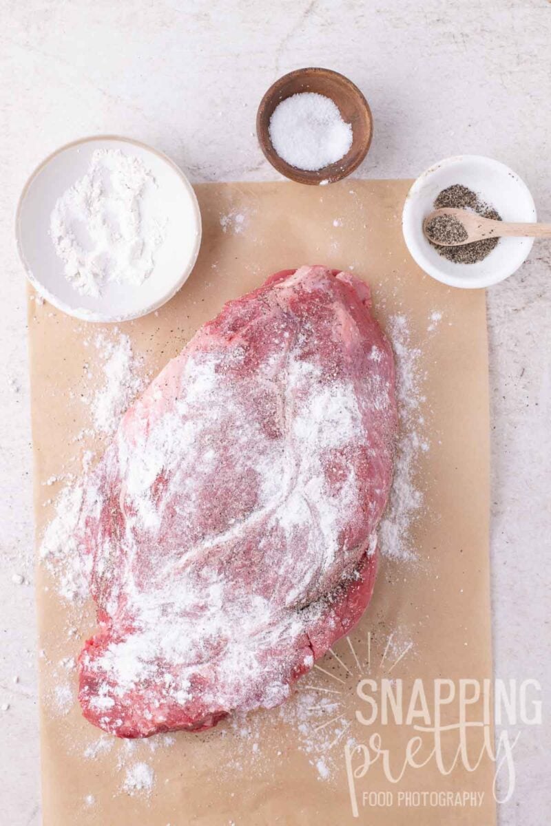 A raw beef roast coated in flour and pepper on parchment paper, with small bowls of flour, salt, and pepper nearby.