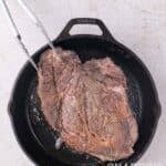 A pair of metal tongs flips a large, seared beef roast in a cast iron skillet.
