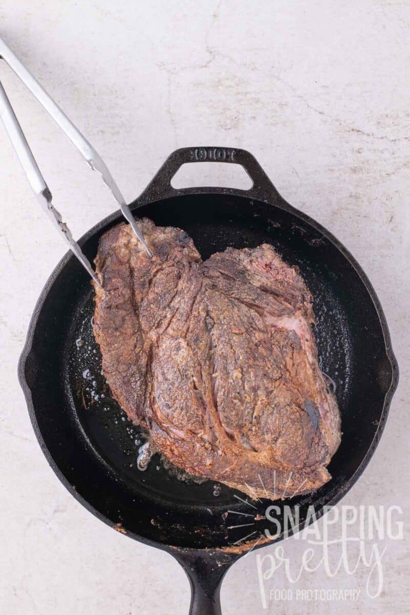 A pair of metal tongs flips a large, seared beef roast in a cast iron skillet.