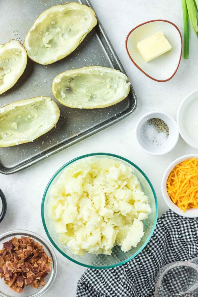Hollowed potato skins on a baking sheet next to a bowl of mashed potatoes and small bowls of bacon, cheese, butter, and seasonings.