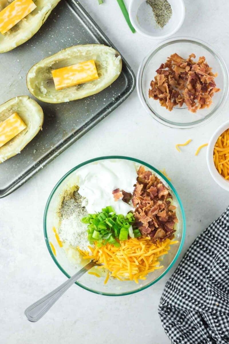 Bowl of potato filling ingredients including sour cream, bacon, green onions, and shredded cheese, with two halved potatoes on a tray nearby.