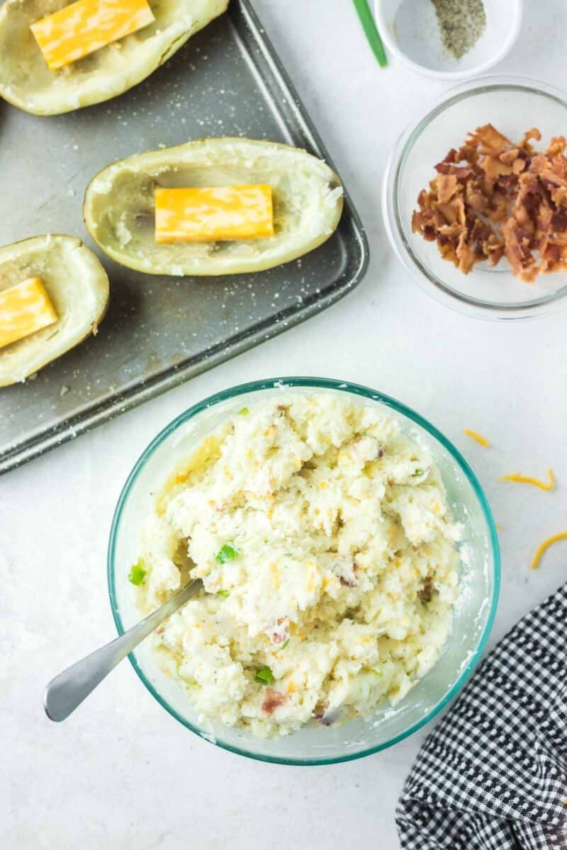 A glass bowl filled with potato mixture, a baking sheet with potato skins and cheddar cheese slices, and a small bowl of crumbled bacon.