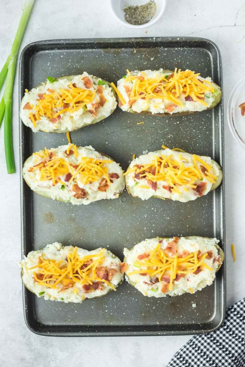Six potato skins topped with mashed potato filling, shredded cheddar cheese, and bacon bits on a baking sheet.